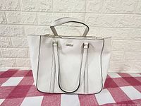 Front view of large white leather Kate Spade purse showing straps and structured design against checkered fabric background.