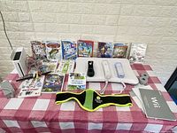 Photo showing Nintendo Wii console and sensor bar with cables, two Wii remotes with wrist straps and silicone covers, Wii Fit balance board, Zumba belt, and 12 Wii game cases lined up on table.