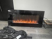 Front view of black glass rectangular electric fireplace heater on floor, flame effect illuminated, remote and bracket beside.