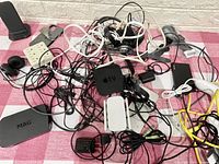 Photo showing an assortment of tech wires, charging cables, an Apple TV box, MAG box, power adapters, and earphones tangled together on a pink checkered cloth.
