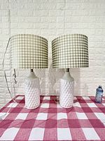 Two white ceramic table lamps with textured diamond pattern on the base and drum lampshades with tan polka dots placed on a red and white checkered surface against a brick wall.
