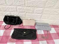 All four clutches and wallet displayed on a pink and white checkered cloth: black crescent handle clutch, beige Tory Burch wallet, silver beaded small clutch, black floral embossed clutch.