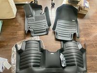 Three black contoured floor mats and a plastic bag of mounting hardware displayed on wood floor