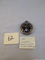 Top-down photo of the 1950s Westclox Pocket Ben pocket watch with black dial and gold numerals. Paper note indicating the watch details is beside it.