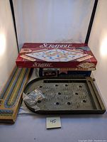 Lot showing boxed Scrabble game on top, two wooden cribbage boards on the side, wooden tabletop pinball game at the bottom with metal pins and a small ball launcher, and a bag of small parts.