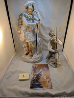 Two ceramic Don Quixote figures photographed with a Lladro brochure; features the larger Staffordshire figure with colorful plaid cape and a smaller kneeling Lladro figure holding a sword.