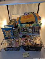 Group view of two tins, deck box, cards laid out, dice and energy cards, jumbo card and poster behind