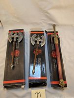 Three miniature weapon replicas displayed on black and red Denix packaging: two double-sided axes and one long sword with sheath.
