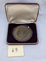 Bronze medallion inside its case on purple velvet with white satin interior marked 'Raymond & Fils Ltée Montreal'.