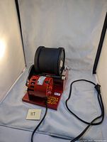 Front angled view of the rock tumbler showing the red metal base, black rubber barrel and attached electric motor with power cord.