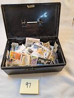 Open metal tin box showing a large quantity of assorted postage stamps inside, across different countries and years, both used and unused.