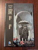 Front cover of DC Comics Batman Earth One graphic novel showing Batman and two characters with flowers in foreground and rain