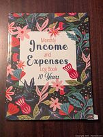 Front cover of a monthly income and expenses log book with decorative floral design and title text.