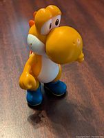 Front-angled view of the orange Yoshi figure standing on wooden floor showing nose, eyes, and blue shoes