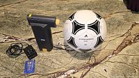 Airman handheld cordless inflator with attached needle nozzle, charger, three silver tone air pump needles and nozzles, and Adidas Tango size 5 football on marble surface.