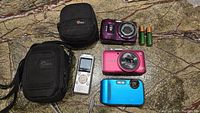 Lot contents showing three digital cameras in blue, pink, and purple, two black camera cases, silver voice recorder, and two AA rechargeable batteries all arranged on marble surface.