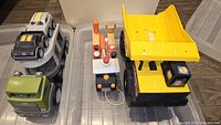All three vehicles of the set visible from above: a green plastic truck carrying two toy cars, wooden truck with tools, and large yellow dump truck.