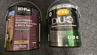 Two paint cans side by side: Behr Premium gray Elastomeric Masonry, Stucco & Brick Paint and CIL DUO semi-gloss red paint.