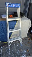 Used 3 step aluminum ladder with blue top platform, showing signs of heavy use, placed outdoors near a covered object.
