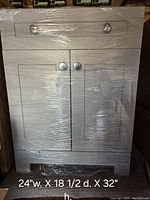 Front view of gray bathroom sink cabinet wrapped in plastic film, showing top drawer, cabinet doors, and chrome knobs with size details overlay.