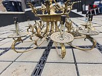 Close-up angled view showing eight brass arms with intricate leaf and floral designs, each supporting a candle-style socket for light bulbs. Visible patina and wear consistent with age and brass construction.