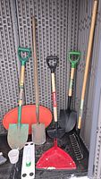 Full view showing four shovels with wooden handles and plastic grips, a large red dustpan, and a black hand rake inside a storage bin