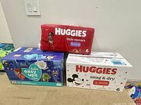 Three packs of pull-up diapers: red Huggies Little Movers size 6, white Huggies Snug & Dry Mickey Mouse size 6, and blue Pampers Easy Ups 5-6, arranged on floor.