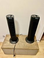 Two standing black tower fans on a wooden box, showing frontal and side views.