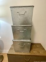 Three large metal storage bins stacked vertically, showing front view, lids on top, each with side handles