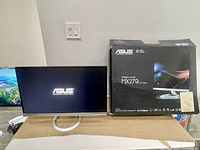 Asus MX279 monitor powered on displaying ASUS logo, next to the original packaging box.
