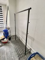Full view of the metal coat rack positioned against a wall on wood floor, showing the hanging bar, hooks, and base shelf.