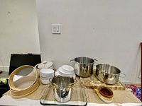 Kitchen items arranged on table showing pans, plates, glass baking dish, wooden cutting board, bamboo steamers, bottle chiller and ceramic vowel
