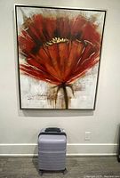 Frontal photo of the large framed floral painting hanging on a white wall with a suitcase below it for size reference.