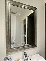 Large rectangular wall mirror mounted on a bathroom wall above a sink area, showing clear reflection and silver textured frame.