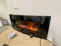 Front view of black linear electric fireplace heater powered on showing simulated flame with orange and blue hues. Remote control is positioned in front of the unit and plastic-wrapped accessories are on the left side.