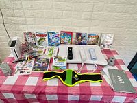 Full overview of Nintendo Wii console, 2 remotes, balance board, sensor bar, cables, Zumba belt, instruction manuals, and 12 game titles.