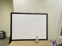 Front view of large whiteboard with black frame, sized 48 x 36, placed against a white wall with a bottle at the base for scale.