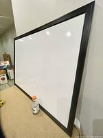 Whiteboard shown diagonally leaning against wall on floor with bottle for scale