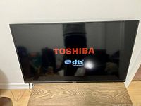Front view of Toshiba flat screen TV powered on showing Toshiba and DTS TruSurround logos on screen.