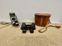 Photo showing side view of the vintage video camera with three lenses and attached strap beside Carl Wetzlar binoculars and leather case on carpeted surface.
