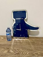 Front view of a blue hand-shaped wall clock branded NeXtime with white clock hands and markers, placed on a wooden floor next to a water bottle for size reference.