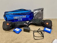 Pair of black Wildyak heated gloves laid out flat with two blue batteries, charger, instruction manual, and blue storage bag behind