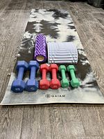 Yoga mat with hand weights and foam rollers arranged on wood floor, showing color and branding of items.