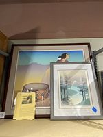Two framed lithographs on a shelf: larger 'Tonto Woman' by R.C. Gorman and smaller 'Green Cove' by James Hagen