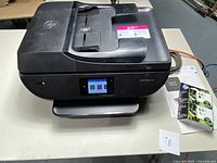 Front view of the HP Envy Photo 7855 printer showing top paper input and closed output tray with ink cartridges beside it.