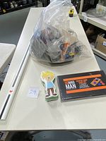 Overall display of lot items including plastic bag with cosplay sword and cloth, a desktop cornhole game box, and a flat cutout figure of a boy on the table.