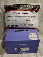 Photo showing the packaged MdBebbron projector screen pouch labeled with 120 inch size, 16:9 scale, and support for 4K, HD, 3D. The AuKing projector box is below it.