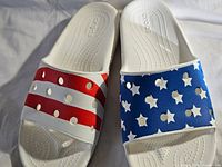 Pair of Crocs Classic Slides showing red and white stripes on the left and blue with white stars on the right slide, resembling American flag design.