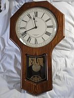 Elgin quartz regulator wall clock front view showing wood finish, clock face with Roman numerals, and pendulum visible behind glass door with "REGULATOR" text.