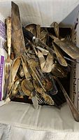 Box filled with assorted tarnished vintage and antique flatware including spoons, forks, knives, and serving utensils, showing various levels of wear.